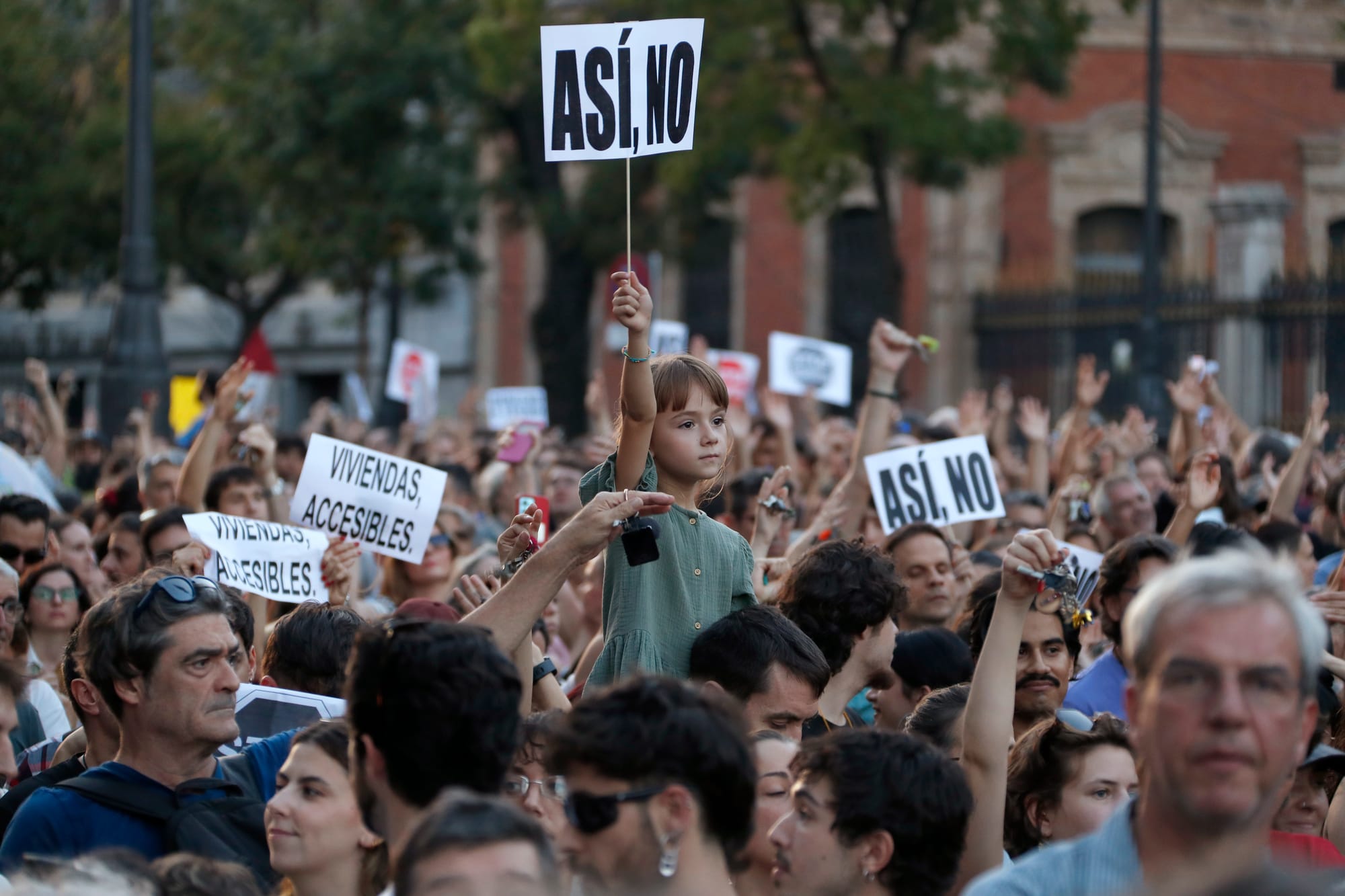 Multitud de personas en una manifestación, destacando en el centro una niña subida a hombros que levanta un cartel blanco con el texto 'ASÍ NO', rodeada de manifestantes con pancartas que piden 'VIVIENDAS ACCESIBLES'.