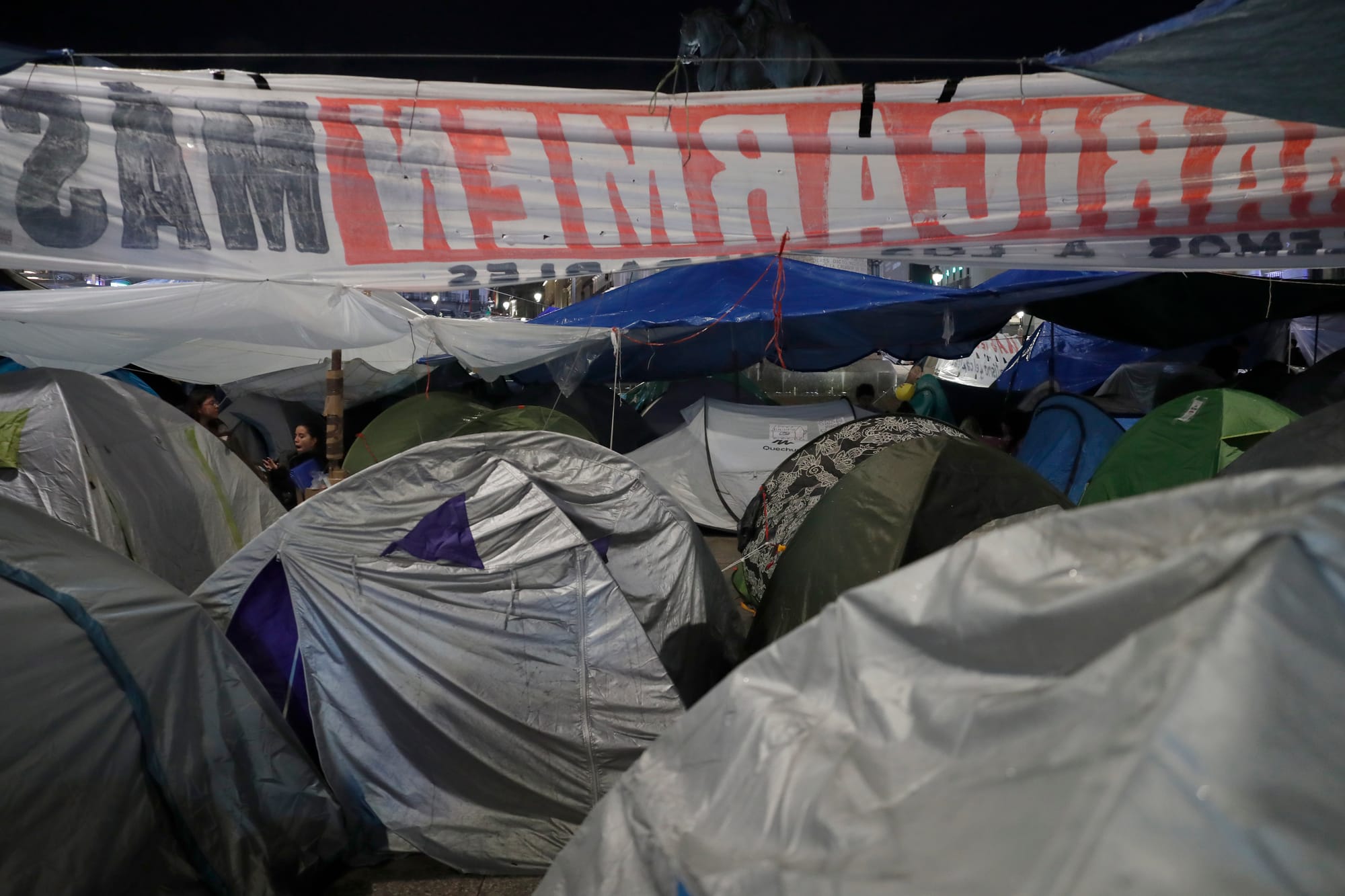 Fotografía nocturna en primer plano de un agrupamiento de tiendas de campaña en la Puerta del Sol de Madrid. Se aprecian varias tiendas de tonos grises y verdes resguardadas bajo plásticos y toldos azules y blancos para protegerse de la lluvia. Sobre ellas, sostenida por cuerdas, cuelga una gran pancarta blanca donde se puede leer a contraluz y del revés el nombre en rojo 'MARICARMEN'.