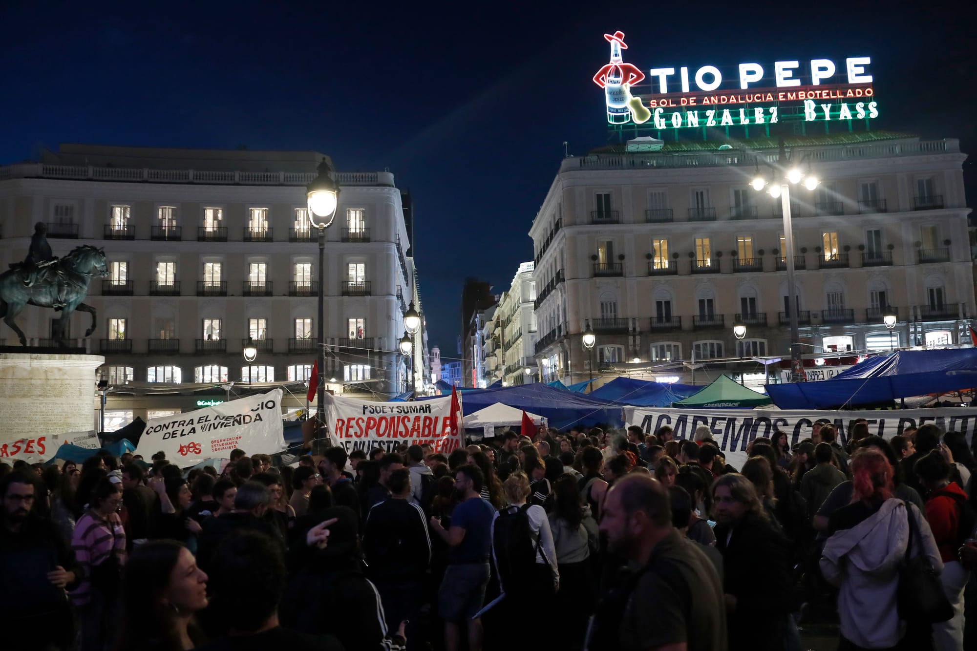 Fotografía nocturna de la multitudinaria acampada ciudadana en la Puerta del Sol de Madrid. Una gran concentración de personas llena la plaza bajo el característico cartel luminoso de Tío Pepe y junto a la estatua ecuestre de Carlos III. Entre la multitud destacan varias carpas azules instaladas al fondo y pancartas reivindicativas con lemas como 'CONTRA EL NEGOCIO DE LA VIVIENDA' y mensajes dirigidos al Gobierno.