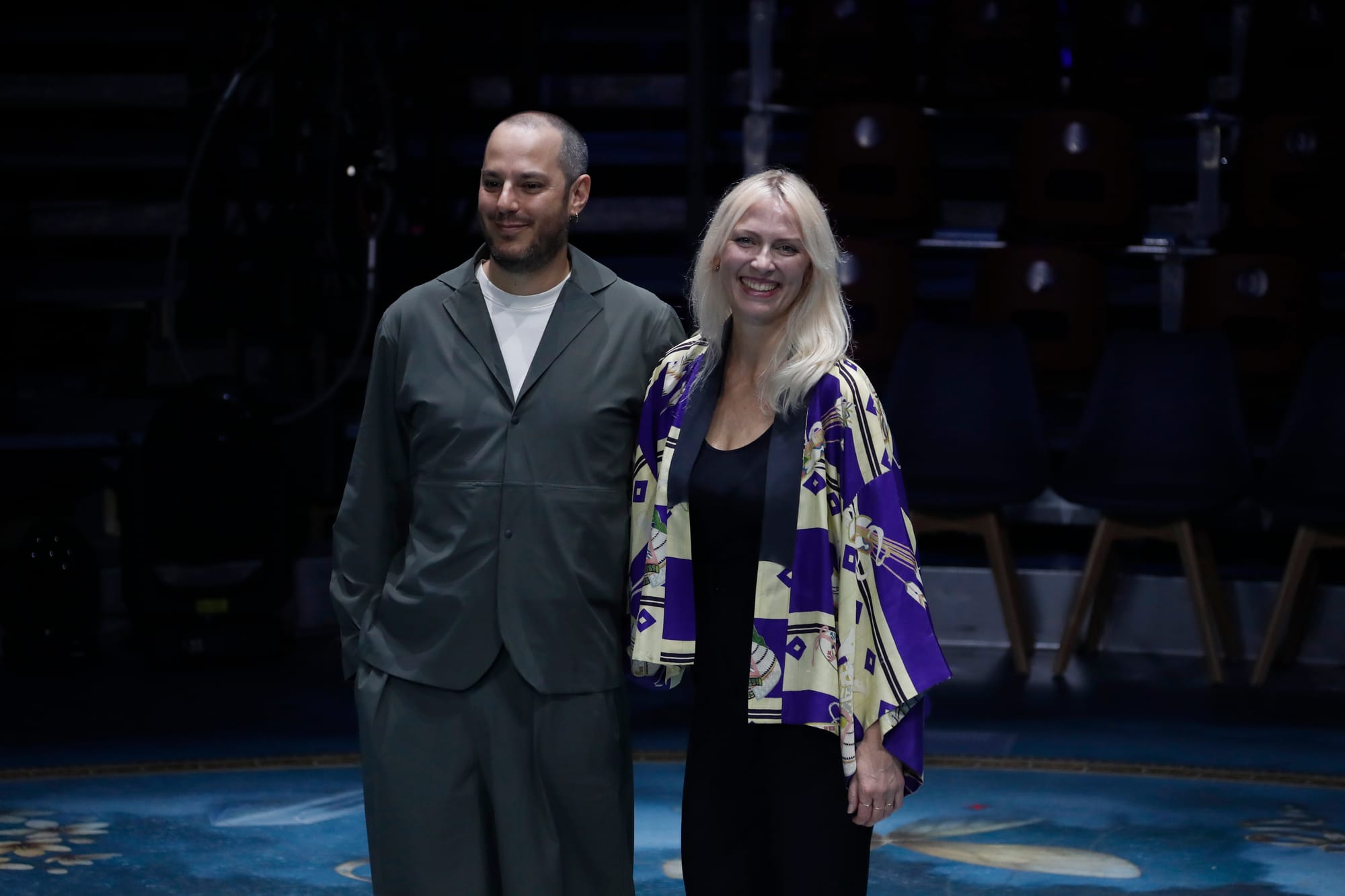 Fotografía de los directores creativos Simone Ferrari y Lulu Helbaek sonriendo ante la cámara sobre el escenario. Simone viste un conjunto gris de corte holgado a la izquierda y Lulu una bata kimonos estampada en tonos morados y amarillos sobre ropa negra a la derecha.