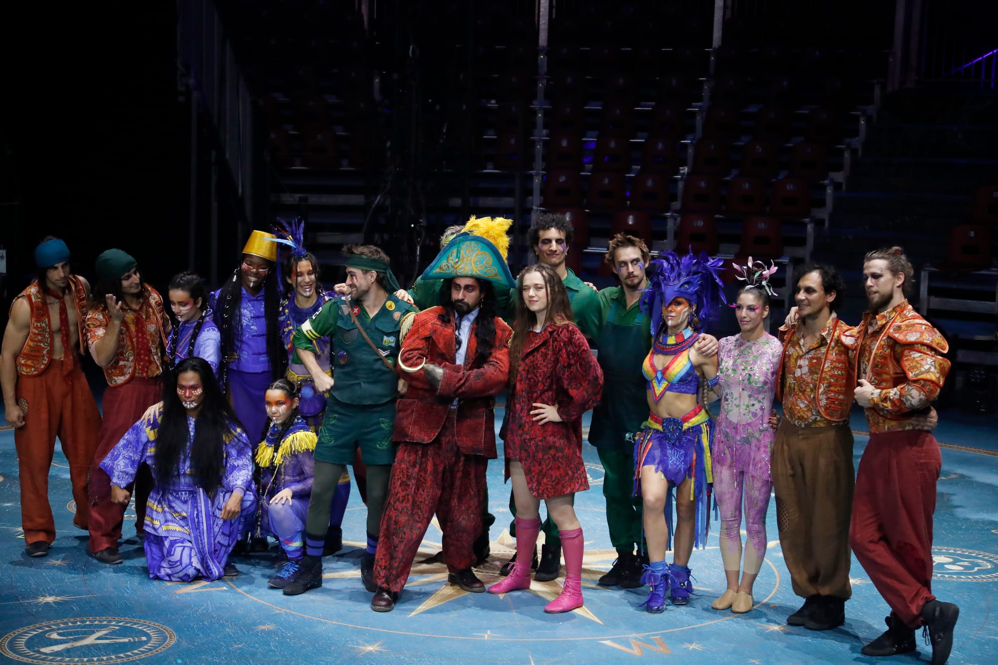 Fotografía de grupo con el reparto completo de 'The Island of Neverland' posando sobre una pista circular decorada con una rosa de los vientos. En el centro destacan los actores caracterizados como Peter Pan, el Capitán Garfio y Tempesta, rodeados por acróbatas y bailarines con vestuario de piratas y miembros de las tribus.