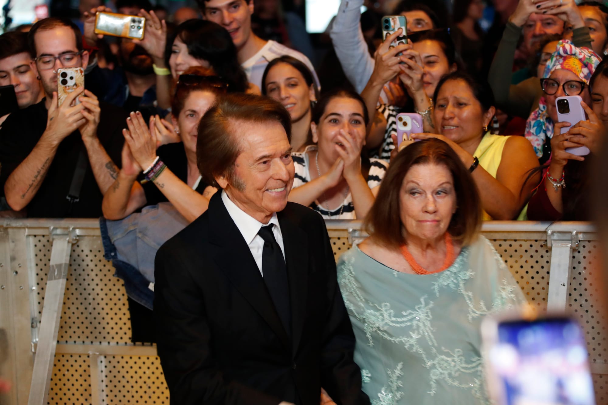 Fotografía del cantante Raphael y su mujer Natalia Figueroa llegando a un evento. Raphael viste traje negro y corbata, y Natalia una blusa verde agua con transparencias y un collar naranja. A sus espaldas, separados por una valla metálica, un gran grupo de admiradores aplauden y les hacen fotos con sus teléfonos móviles.