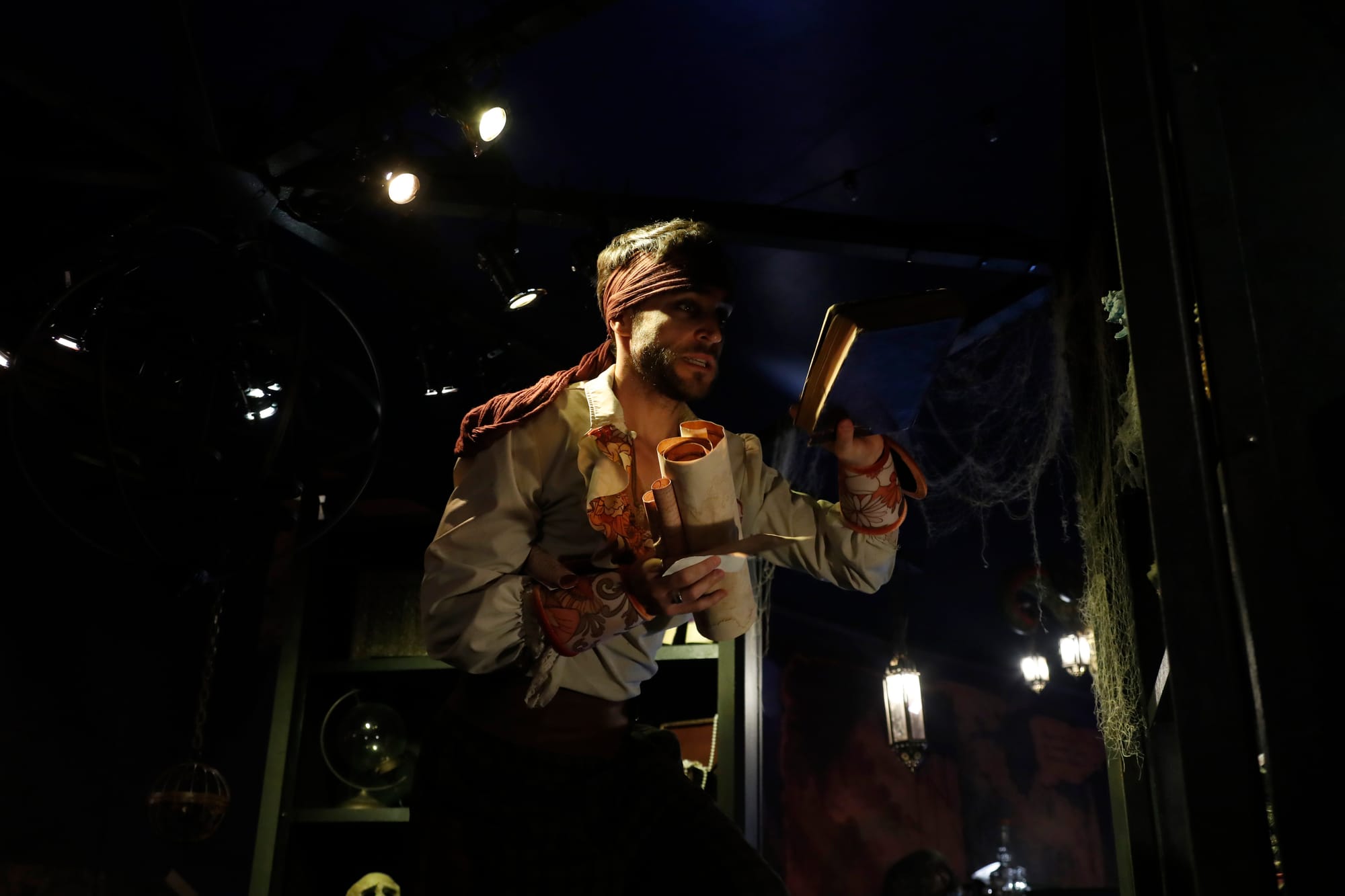Fotografía de un actor caracterizado de pirata con pañuelo en la cabeza y camisa de época, sosteniendo varios pergaminos en una estancia temática de ambientación marina iluminada por focos tenues.