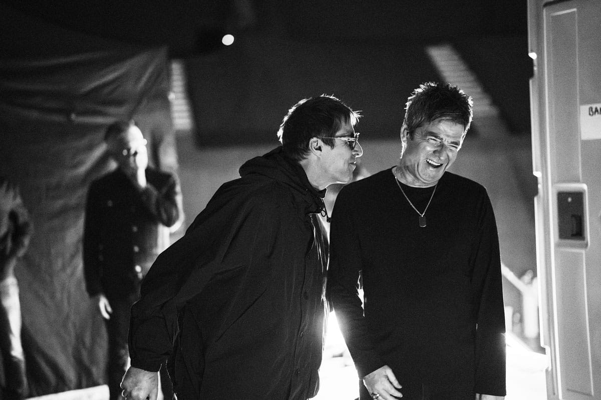 Fotografía en blanco y negro de estilo documental donde se ve a los hermanos Gallagher en actitud relajada y compartiendo risas. Liam, a la izquierda con su característica parka oscura y gafas, se inclina hacia su hermano Noel, quien ríe abiertamente vestido con un jersey oscuro y un collar de chapa militar. De fondo se intuye una figura desenfocada en lo que parece un backstage.