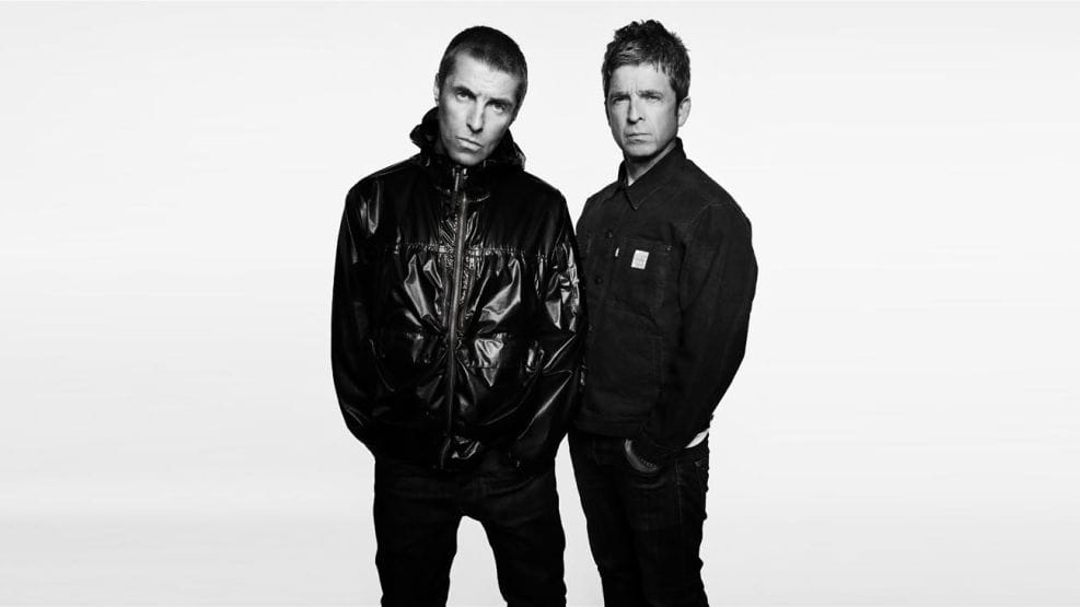 Retrato fotográfico oficial en blanco y negro de los hermanos Liam y Noel Gallagher mirando fijamente a cámara con semblante serio. Liam, a la izquierda, viste una parka oscura abrochada, y Noel, a la derecha, lleva una sobrecamisa oscura, ambos posando muy juntos sobre un fondo claro.