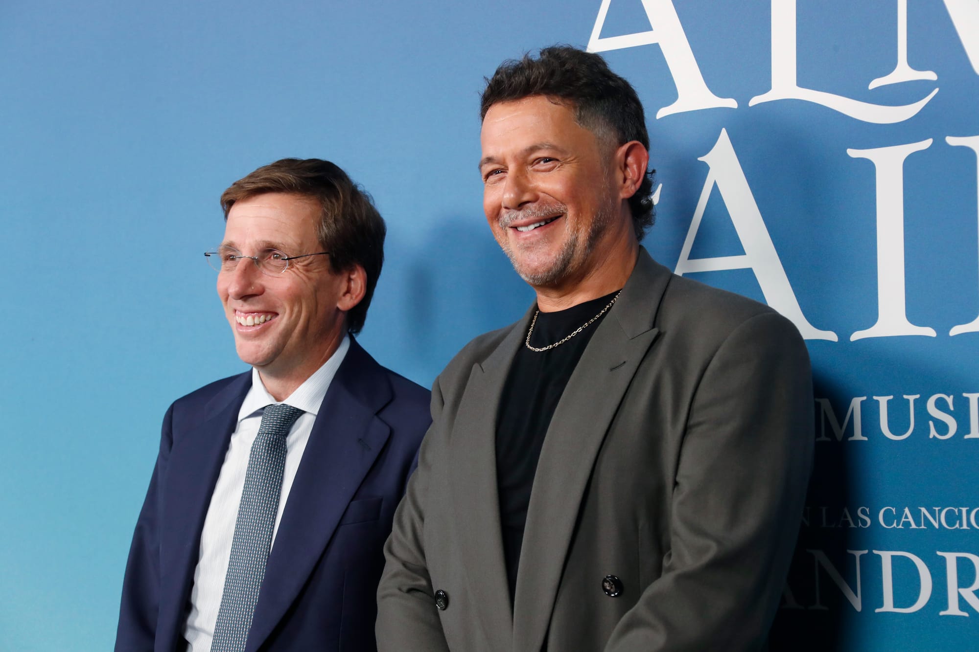 Fotografía en plano medio de José Luis Martínez-Almeida, vistiendo traje azul marino y corbata gris, junto a Alejandro Sanz, con chaqueta gris y camiseta negra con cadena dorada, posando sonrientes ante el photocall azul del espectáculo 'El alma al aire'.