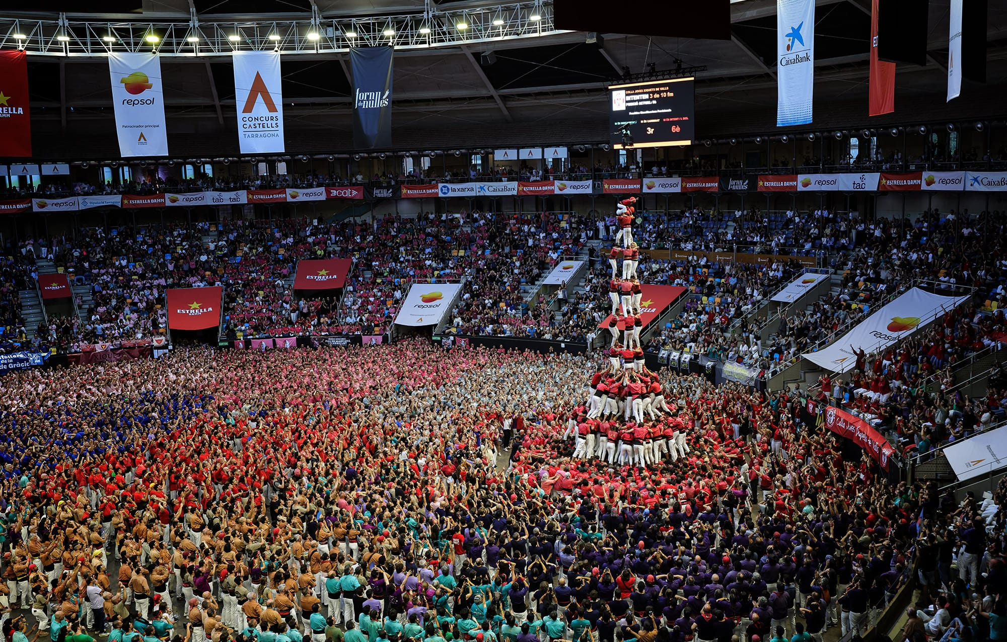 Fotografía panorámica del interior de la Tarraco Arena Plaça donde la Colla Joves Xiquets de Valls, con camisa roja, erige la estructura del 4 de 10 amb folre i manilles rodeada por miles de asistentes que abarrotan la plaza.