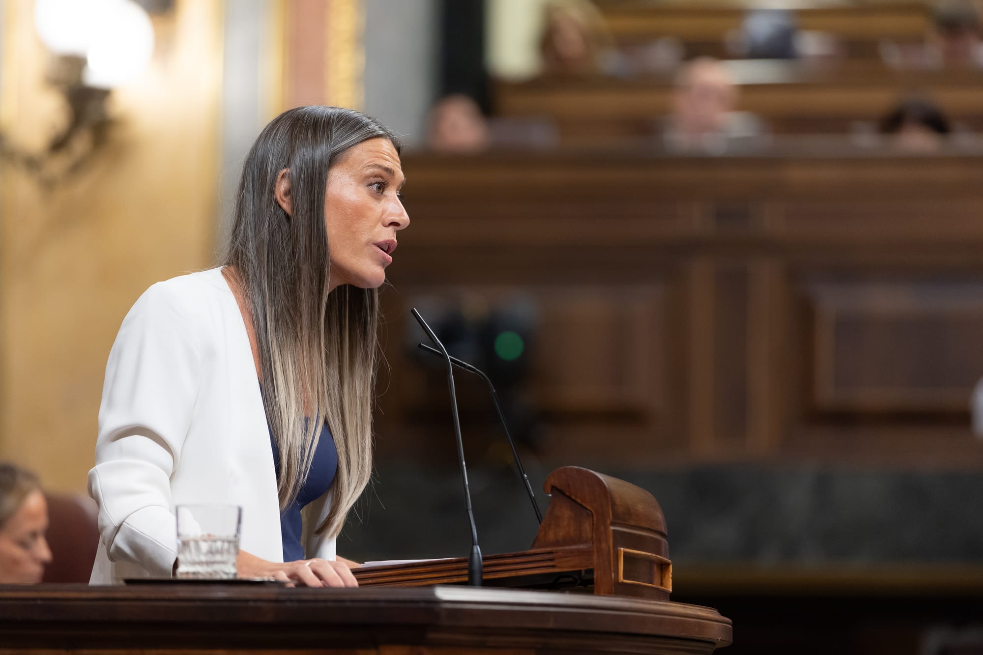 Fotografía en plano medio de perfil de la diputada Míriam Nogueras, vistiendo chaqueta blanca sobre un top azul marino, dirigiéndose al hemiciclo desde la tribuna de oradores del Congreso de los Diputados.