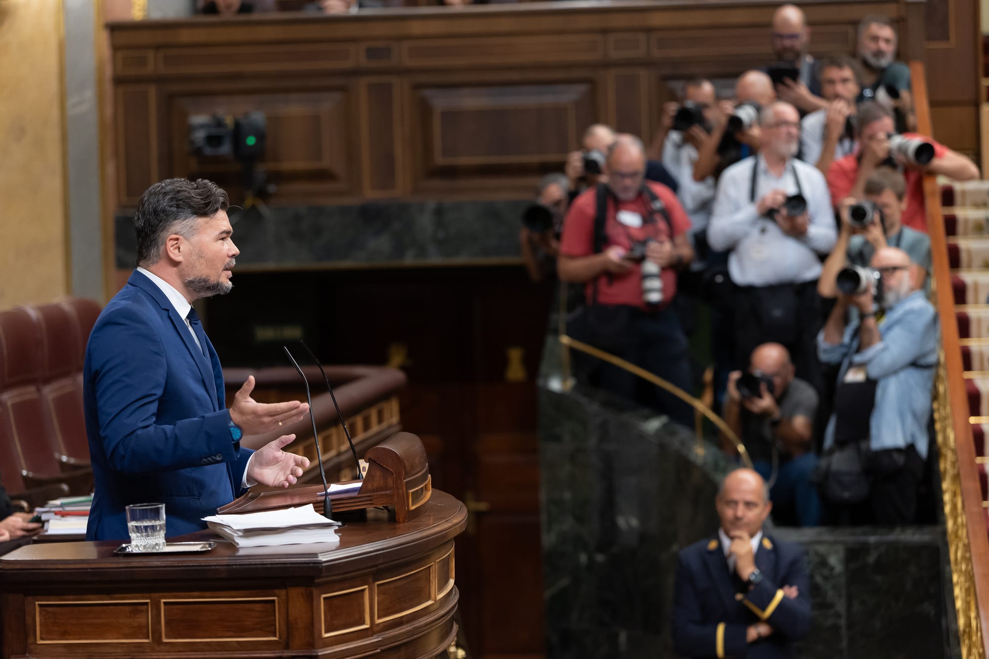 Fotografía en plano medio de Gabriel Rufián, vistiendo traje azul y corbata oscura, gesticulando con las manos abiertas desde la tribuna de oradores del Congreso de los Diputados ante la mirada de reporteros gráficos al fondo.