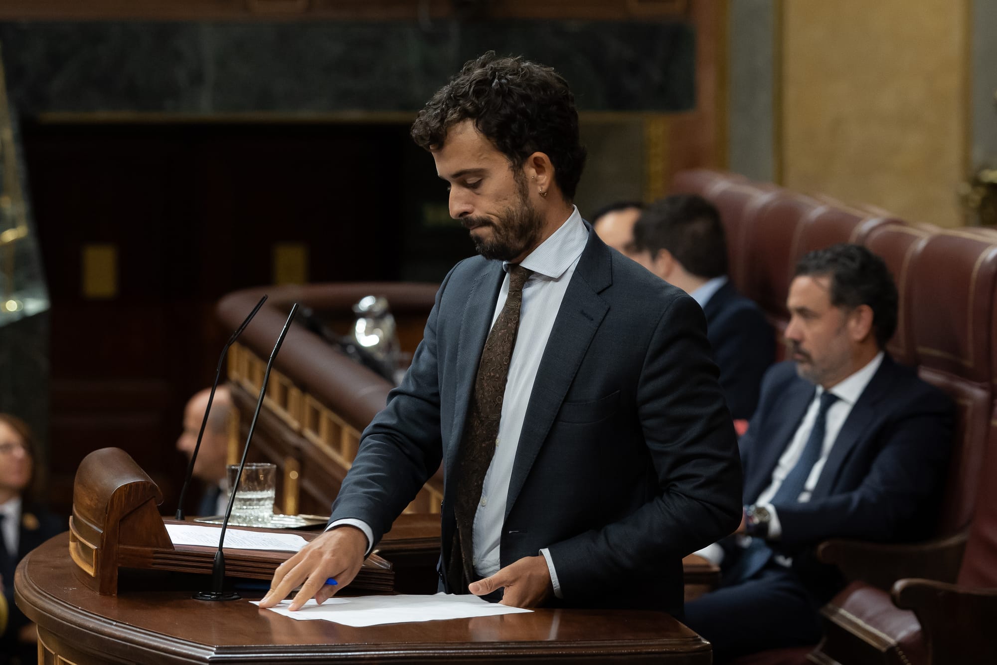 Fotografía en plano medio del diputado Carlos Hernández Quero, vistiendo traje azul oscuro y corbata estampada, dirigiéndose al hemiciclo desde la tribuna de oradores del Congreso de los Diputados.