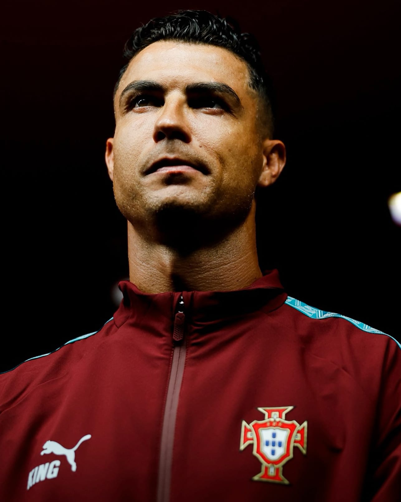 Primer plano vertical de Cristiano Ronaldo con gesto reflexivo y mirada elevada, luciendo la chaqueta roja de entrenamiento de la Selección de Portugal con el escudo de la federación lusa y la marca Puma.