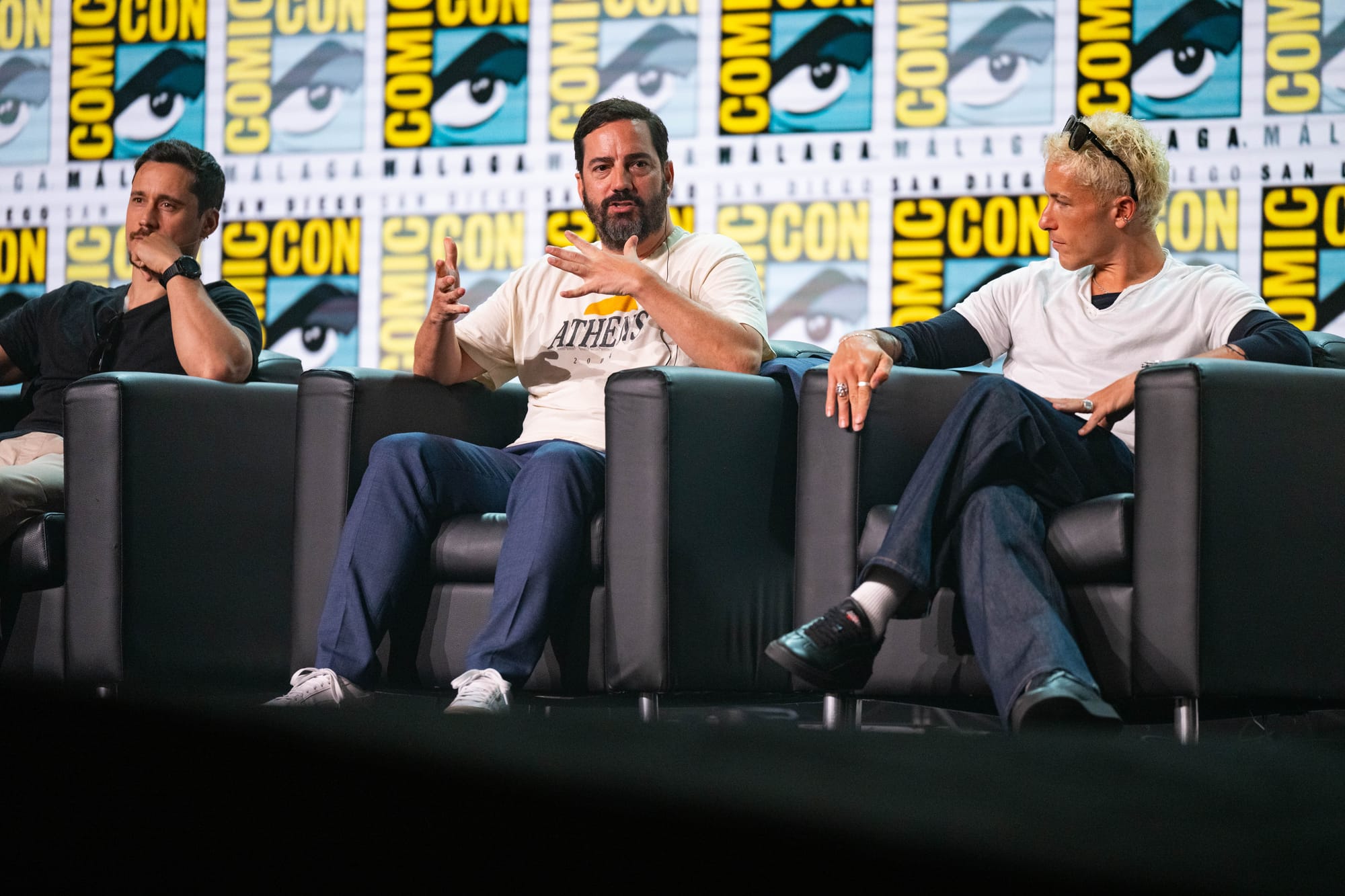 Fotografía en plano medio de tres miembros del equipo de la serie One Piece sentados en sillones negros sobre el escenario: Peter Gadiot a la izquierda apoyando la barbilla en la mano, Lukas Ettlin en el centro gesticulando mientras habla, y Taz Skylar a la derecha escuchando atentamente ante el photocall oficial.
