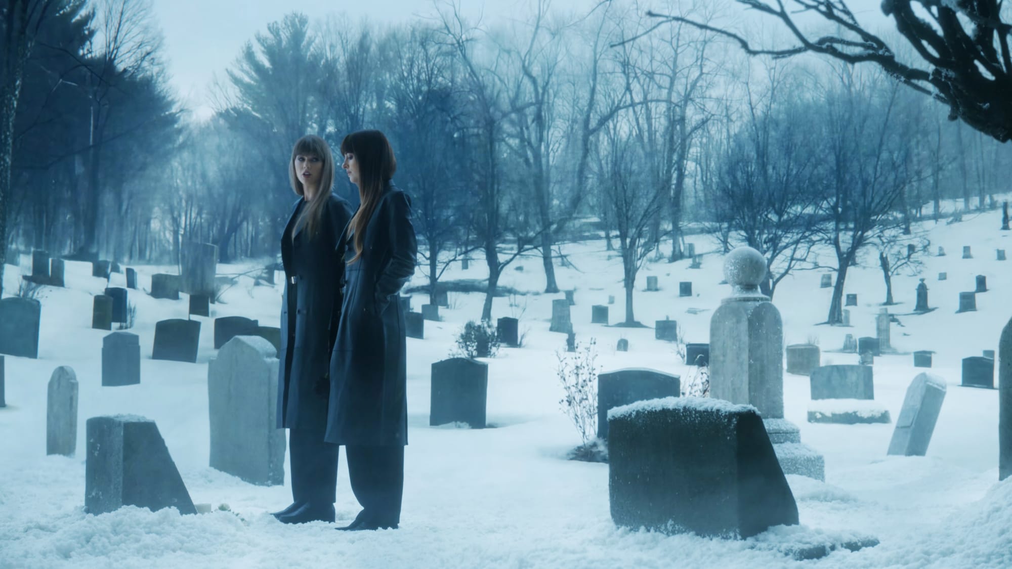 Plano general de Taylor Swift (izquierda) y Dakota Johnson (derecha) luciendo abrigos negros largos de pie en un cementerio nevado repleto de lápidas y árboles invernales, en un fotograma con tonalidad azulada del videoclip 'Patient Zero'.