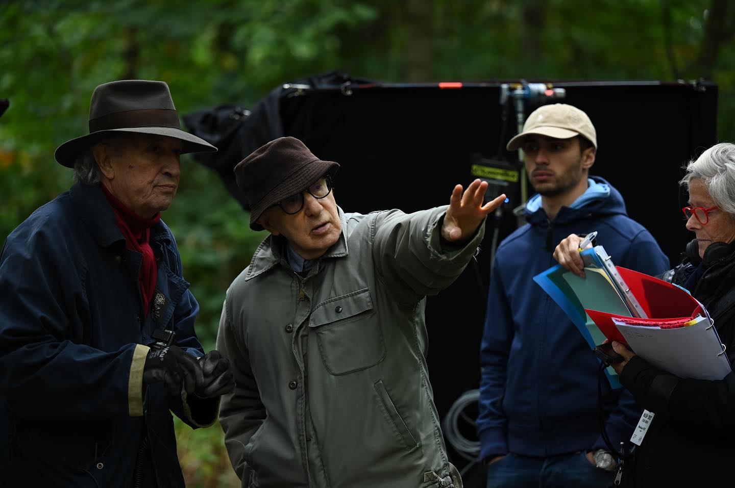 Fotografía en plano medio del director Woody Allen, vistiendo su característico sombrero de tela y chaqueta verde, señalando con la mano izquierda para dirigir una escena en exteriores rodeado por miembros del equipo de producción con carpetas de guion.