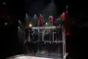 Fotografía de varios acróbatas ejecutando saltos acrobáticos sobre una alta estructura vertical transparente con trampolines en la base. Tres de los acróbatas permanecen en la parte superior.