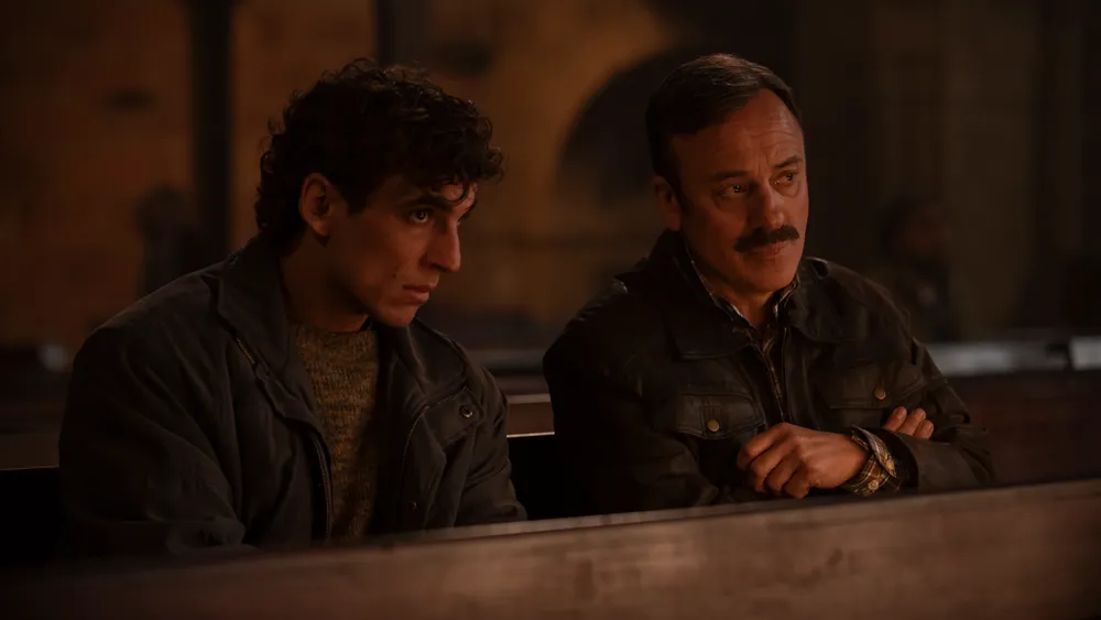 Fotograma de la película 'El Confidente'. Los actores Miguel Herrán, a la izquierda, y Javier Gutiérrez, a la derecha con bigote, aparecen sentados juntos en un banco de madera.