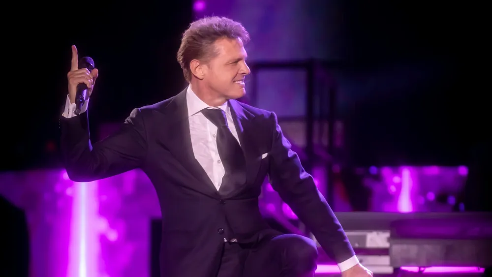 Fotografía en primer plano medio de Luis Miguel actuando en directo sobre un escenario. Viste un elegante traje negro con camisa blanca y corbata oscura.
