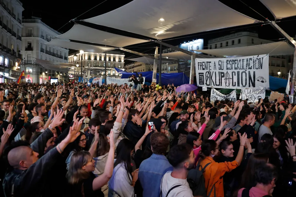 Fotografía nocturna de una gran multitud concentrada en la Puerta del Sol bajo unos toldos blancos tensados. Cientos de personas alzan las manos abiertas al unísono.