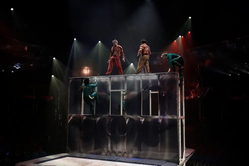 Fotografía de varios acróbatas ejecutando saltos acrobáticos sobre una alta estructura vertical transparente con trampolines en la base. Tres de los acróbatas permanecen en la parte superior.
