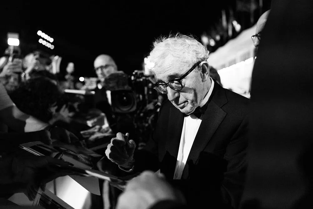 Fotografía en blanco y negro en plano medio del director Woody Allen, luciendo esmoquin con pajarita y gafas de pasta negras, concentrado mientras firma autógrafos a los fans congregados.