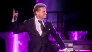 Fotografía en primer plano medio de Luis Miguel actuando en directo sobre un escenario. Viste un elegante traje negro con camisa blanca y corbata oscura.