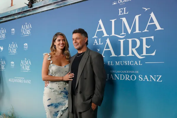 Plano medio de la actriz Stephanie Cayo, luciendo un vestido blanco de tirantes con volantes y estampado azul, junto a Alejandro Sanz, vestuario con chaqueta gris y camiseta negra.